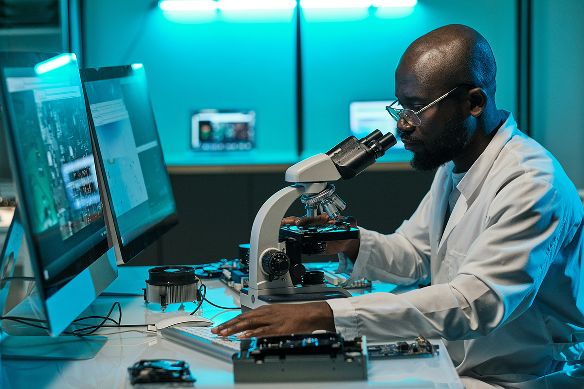 Health researcher looks through a microscope in front of computer monitors.